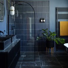 A dark navy ensuite bathroom with rectangular wall tiling, glass arch-shaped shower door, black metal heated towel rail with mustard yellow chevron patterned towel, circular mirror and white bathtub