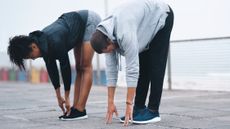 A man and a woman in workout clothes stand outside performing a forward fold. They are hinging at the hips to bend forward, with their arms held straight and fingers touching the floor. They stand on concrete slabs; behind them is a metal railing.