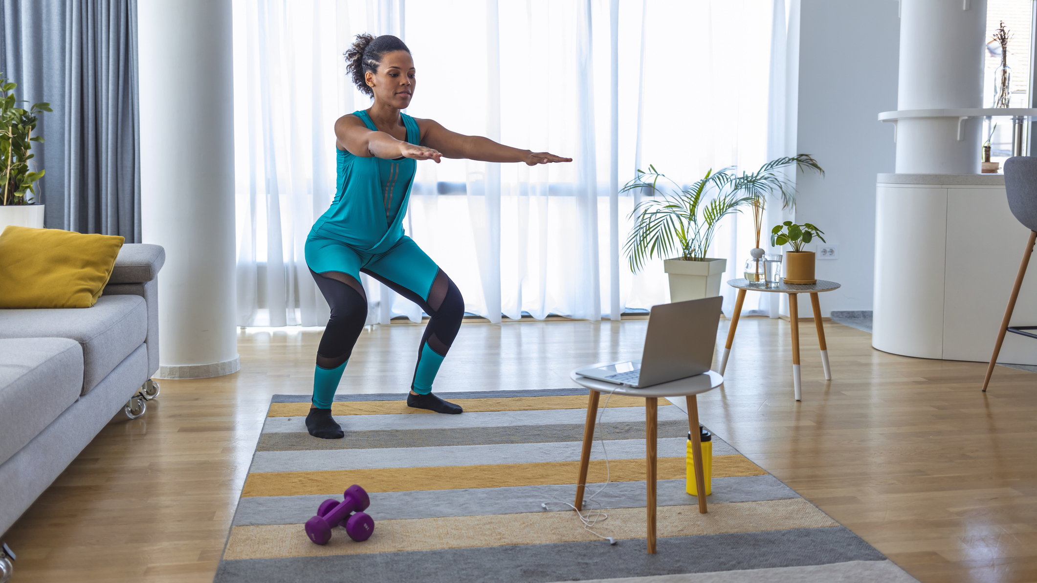 Woman performing the squat exercise in a living room in front of a laptop on a stool