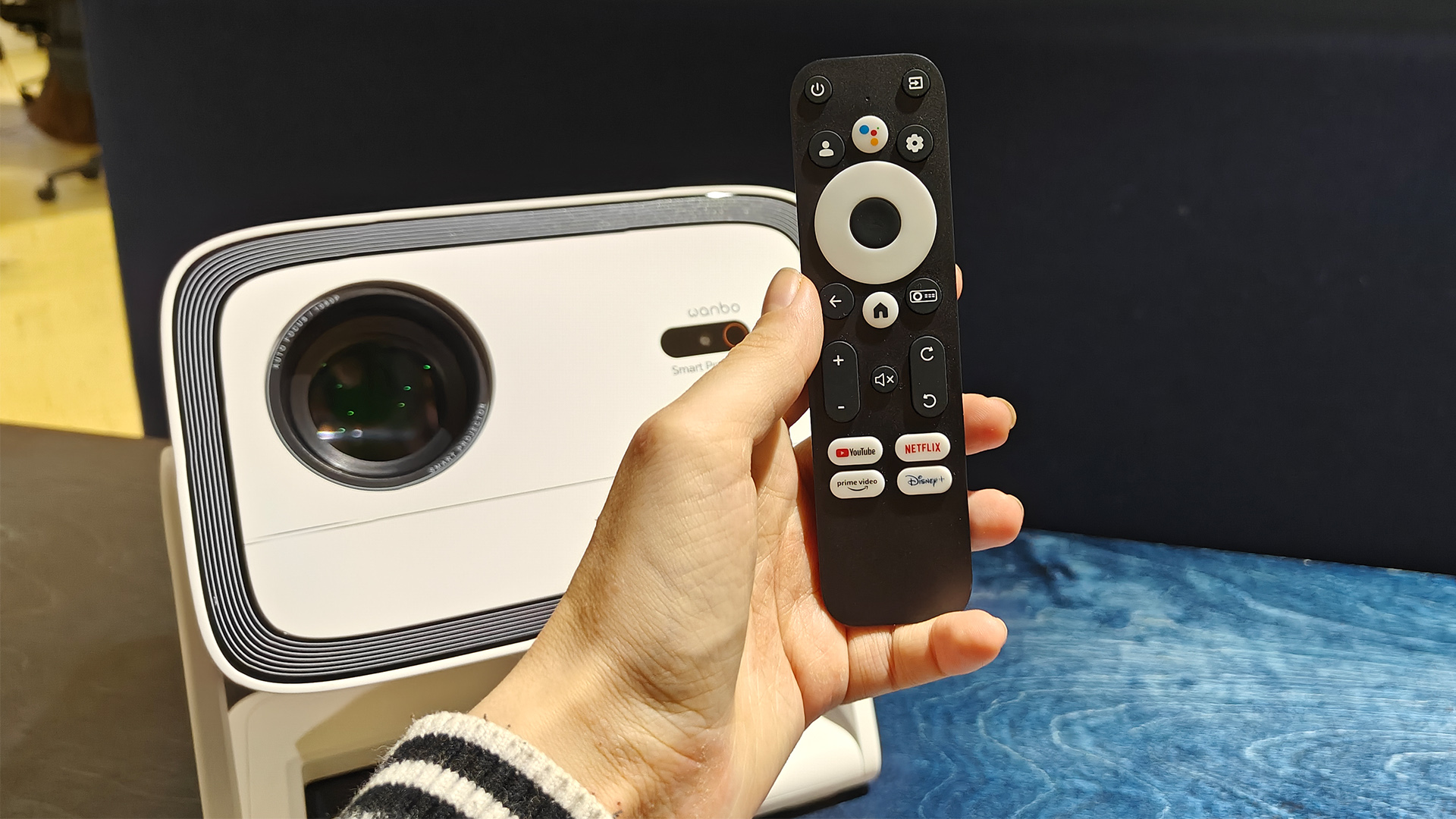Wanbo Vali 1 home cinema projector with remote control held in hand in front of it