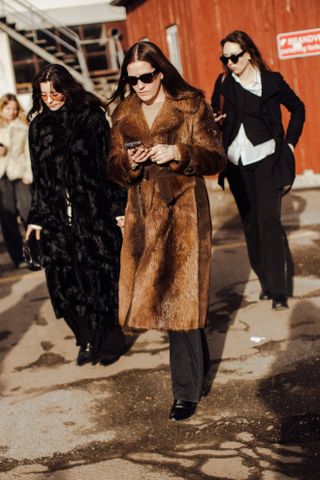 a woman walks down the street at Copenhagen Fashion Week wearing a brown faux fur coat and dark pants