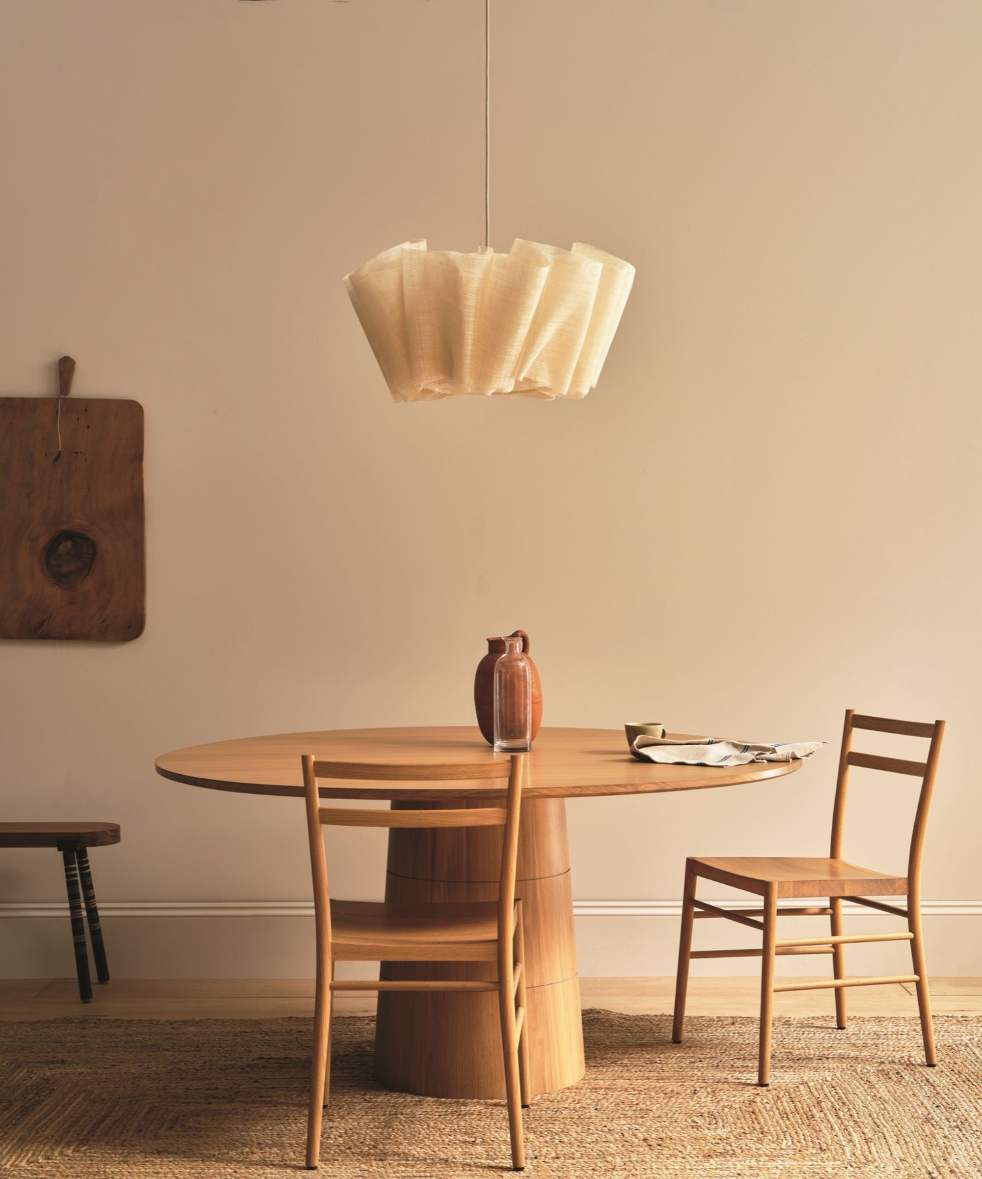round dining table with a woven lamp hanging above with two dining chairs, and a cutting board hanging on the wall