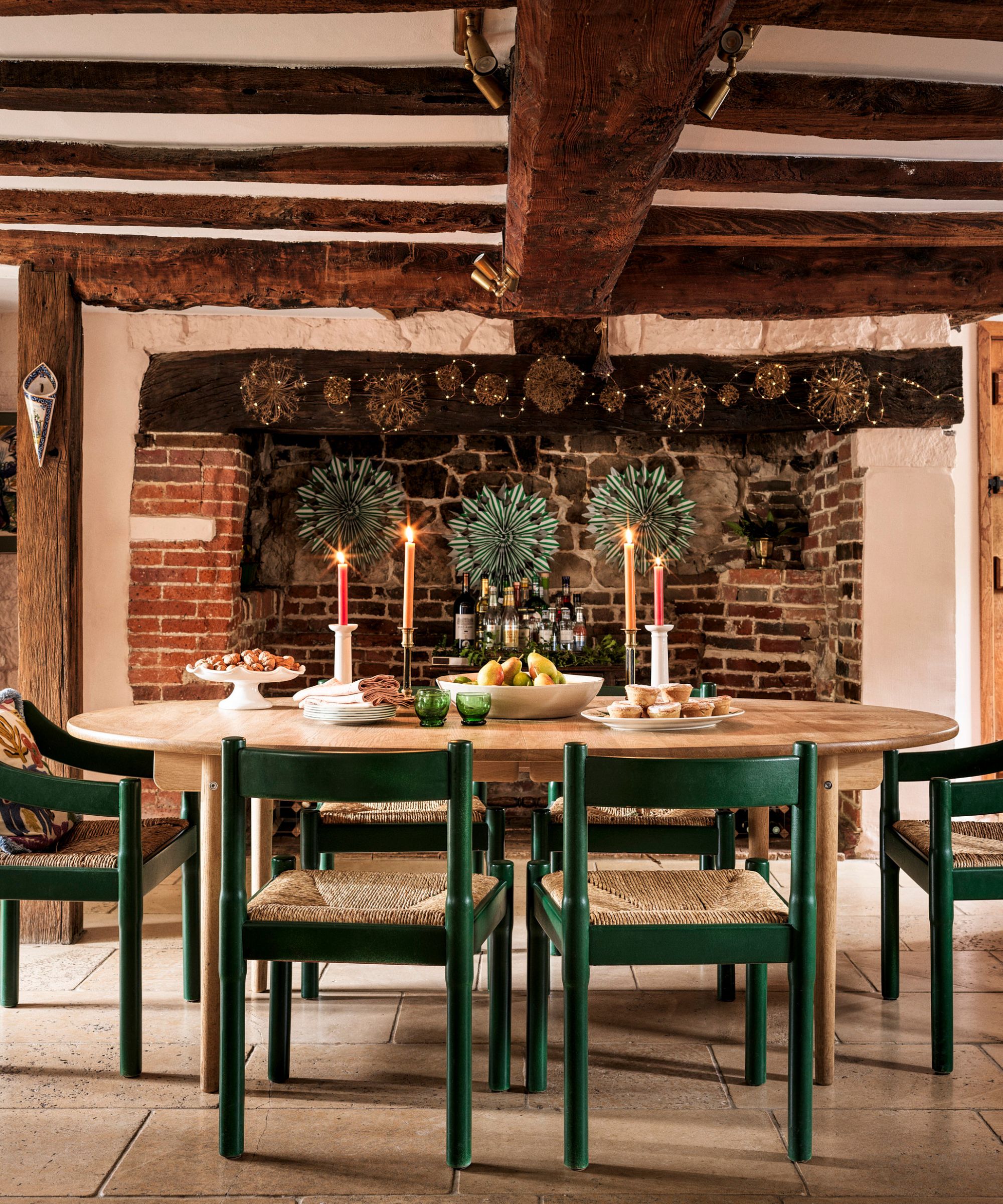 Dining room with wooden beamsm a oval wooden table and green chairs. The is a fireplace behind the table that's decorated with green paper christmas decorations