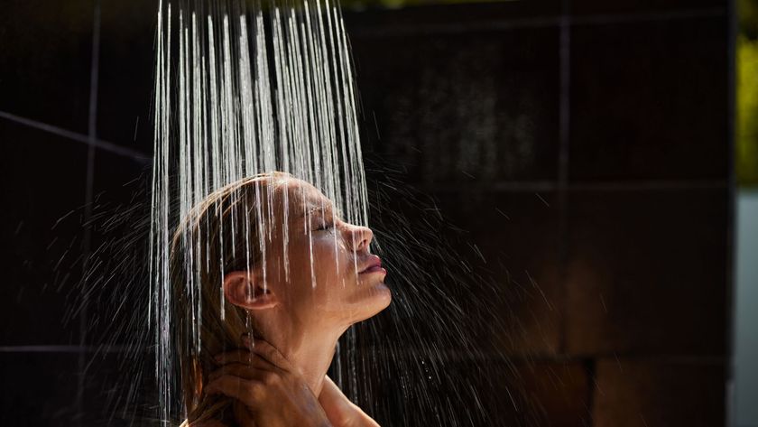 A woman stands in a shower in a bathroom with dark colored tiles on the wall.