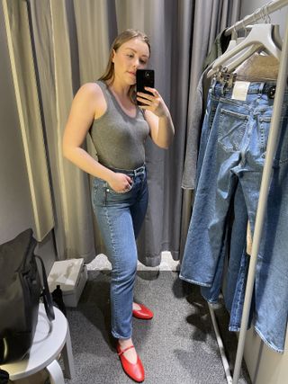 Woman in dressing room wears grey top, blue jeans and red shoes
