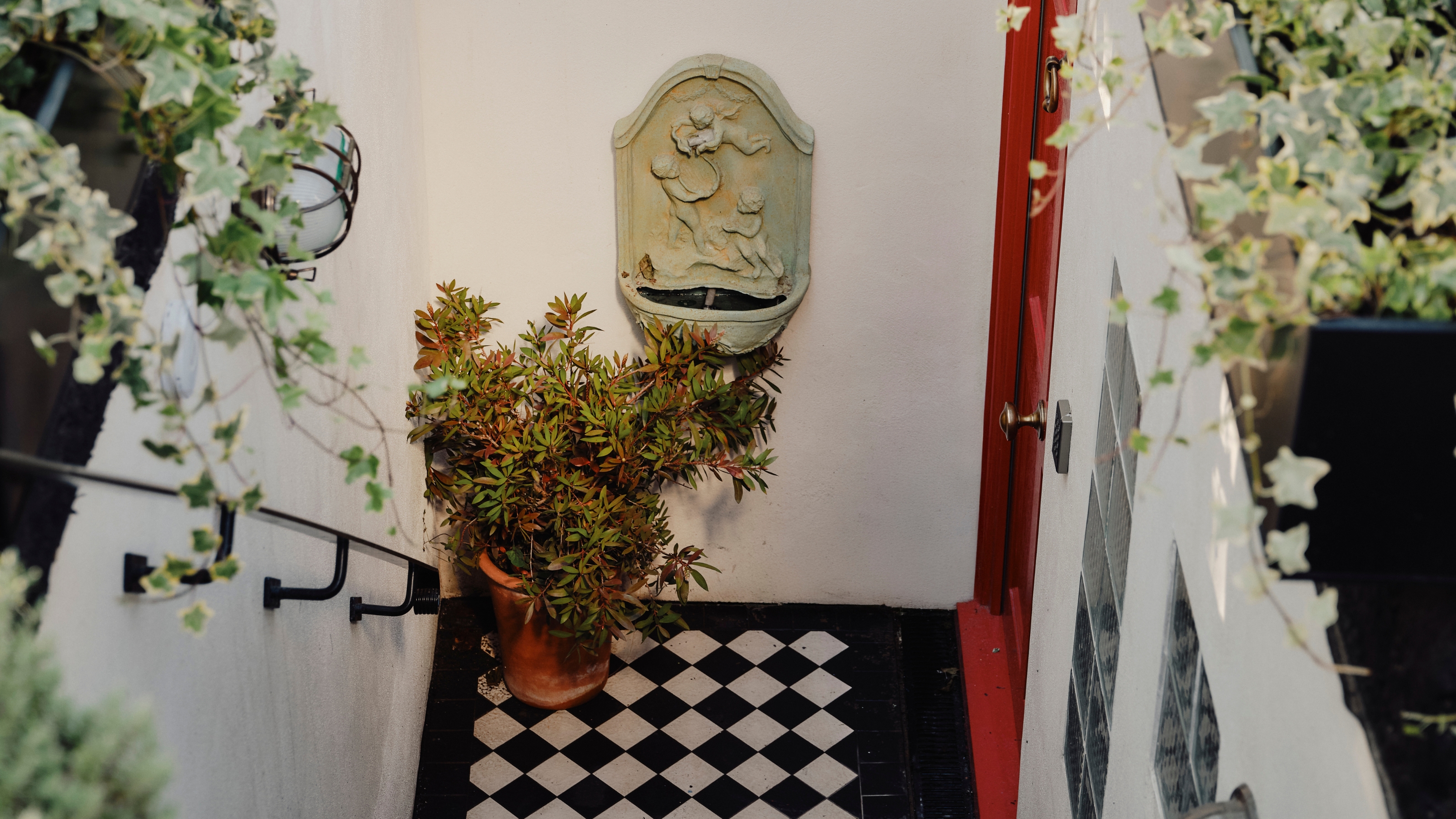 A checkered hallway at the bottom of a staircase is decorated with trailing ivy and potted plants, black and white tiles, a red-painted door, and a marble fountain hanging on the wall.