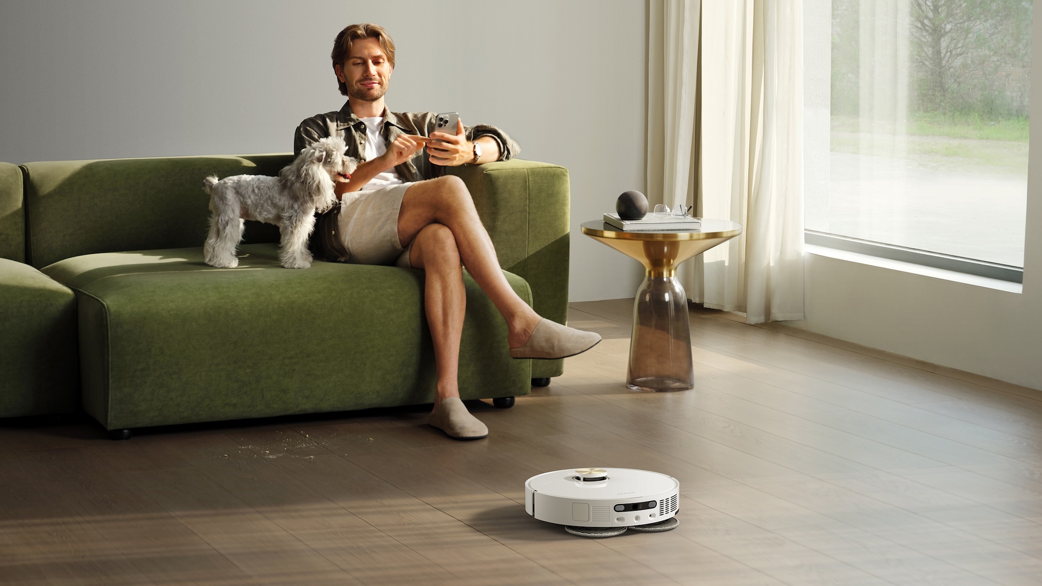 A man and a dog on a green sofa while a Dreame L50 Ultra AE robot vacuum cleans the wooden floor in front of them