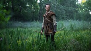 Jack Patten as Robin Hood holding a bow in his hand as he stands in the forest.
