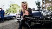 Portrait of American Rock musician Lita Ford as she poses with guitar and a Corvette sports car at a gas station, Chicago, Illinois, September 30, 1984