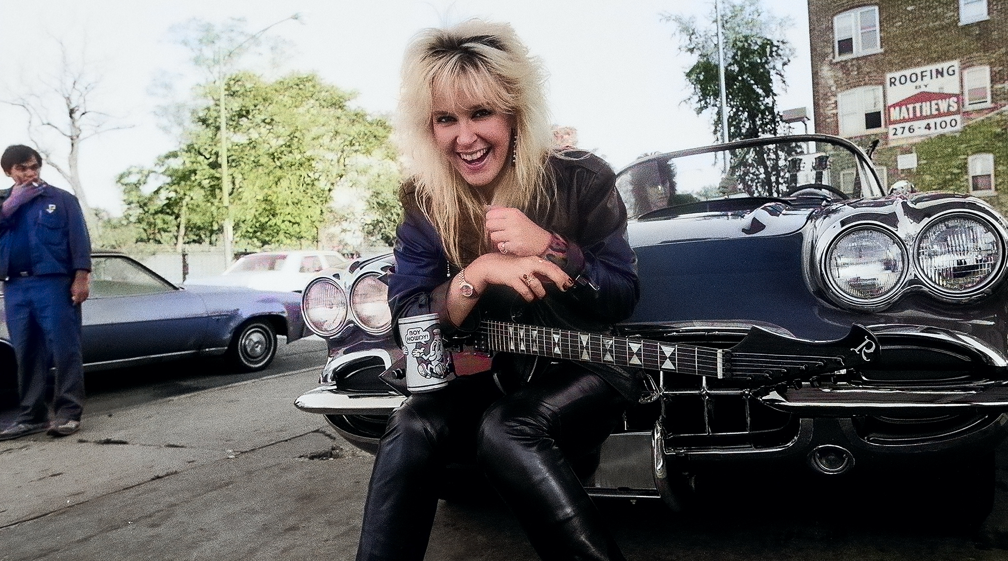 Portrait of American Rock musician Lita Ford as she poses with guitar and a Corvette sports car at a gas station, Chicago, Illinois, September 30, 1984 