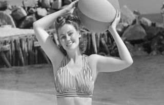 Black and white photo of a woman in a bikini holding a beach ball on the beach