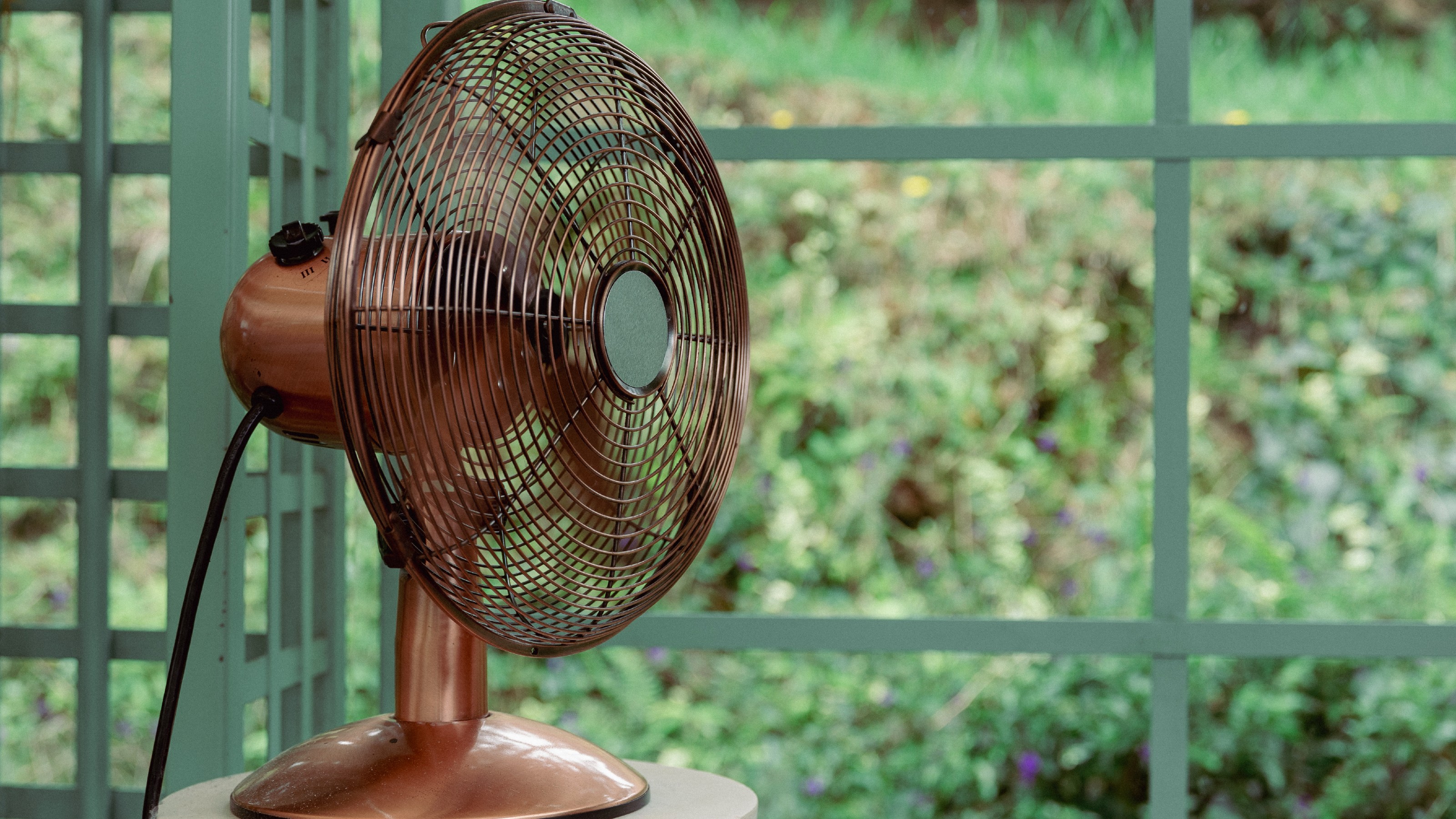 Copper desk fan on a stand in front of green trellis with views to garden greenery