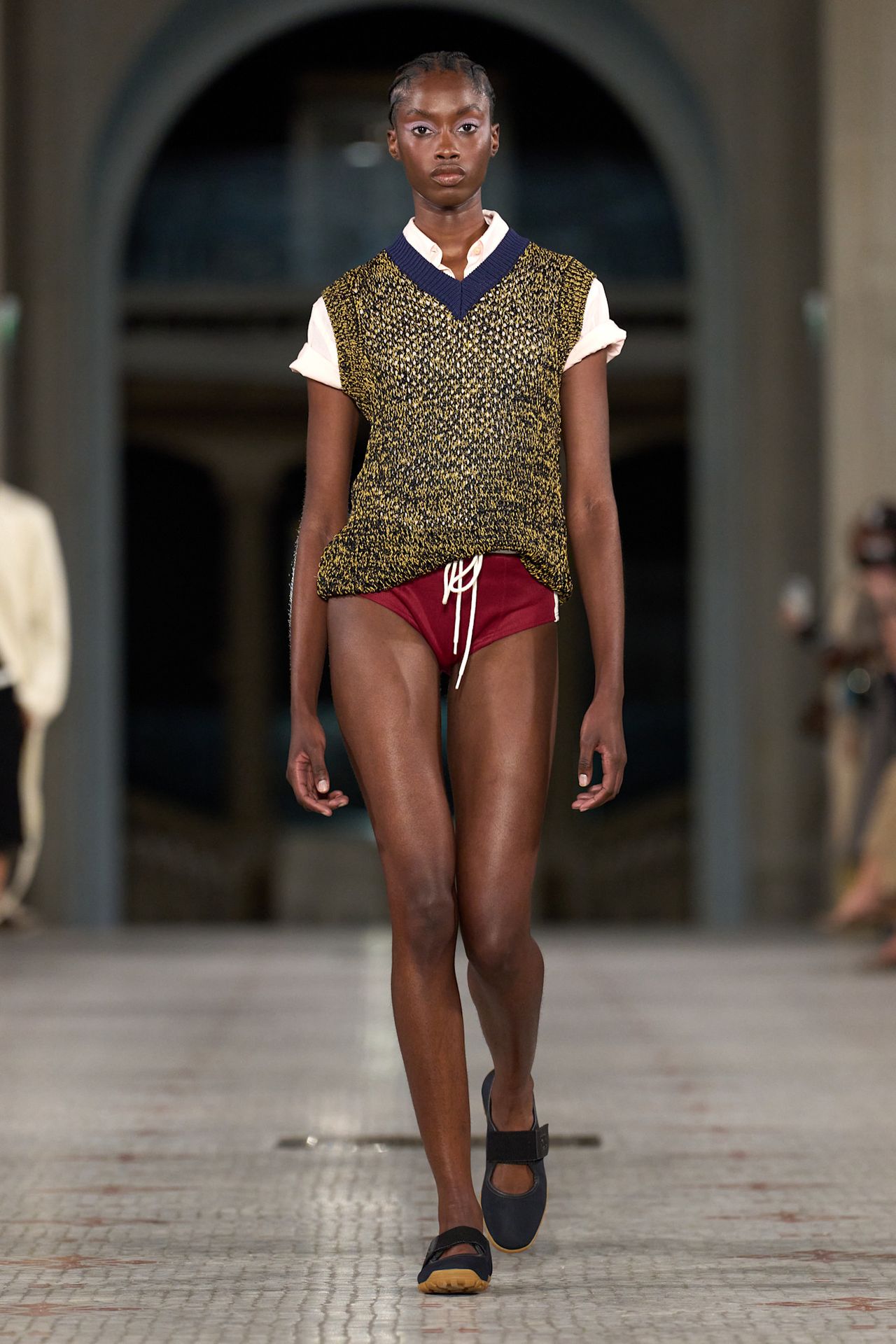 A S/S 25 Wales Bonner model wearing a polo, sweater vest, and mini swim shorts with ballerina sneakers.