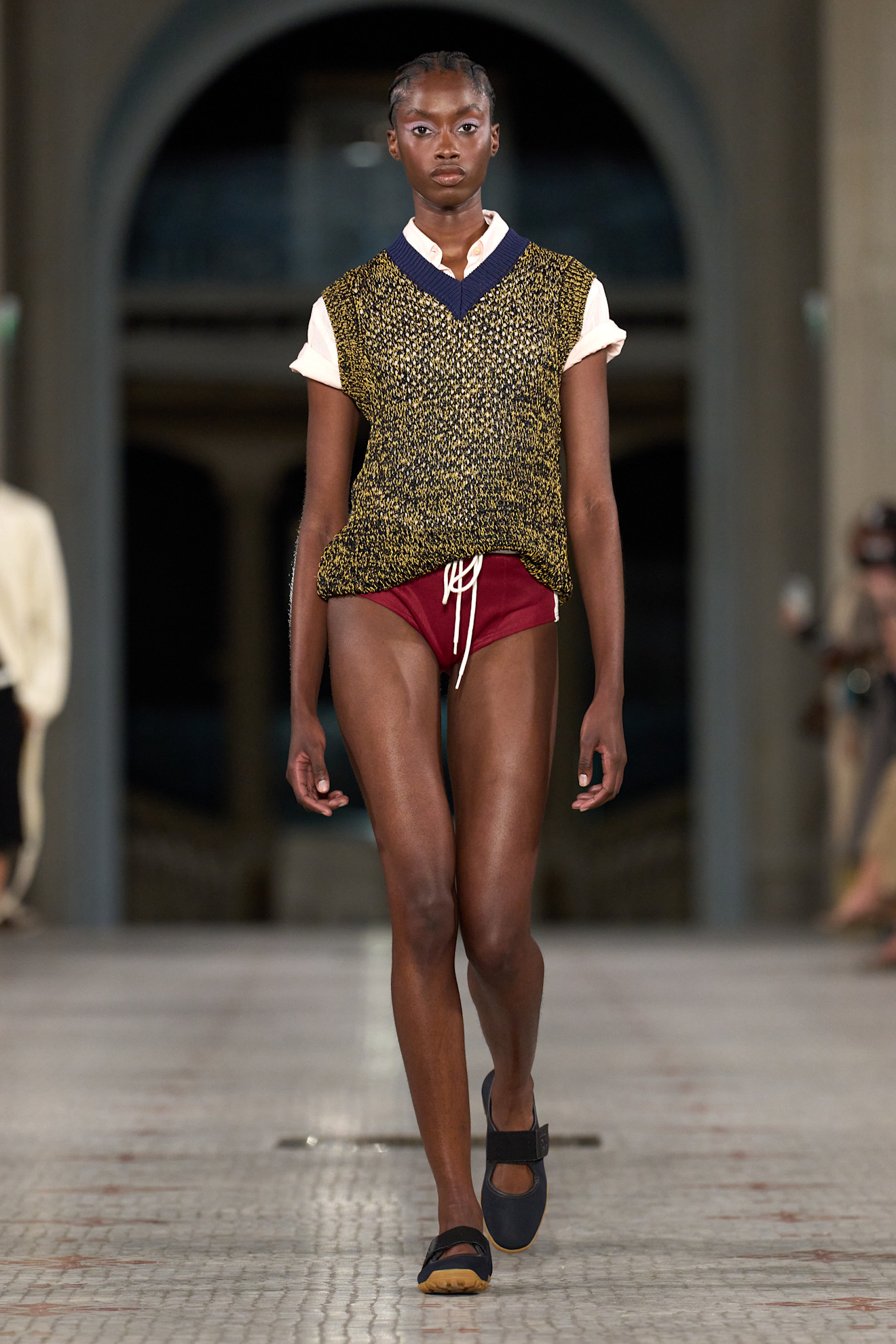 A S/S 25 Wales Bonner model wearing a polo, sweater vest, and mini swim shorts with ballerina sneakers.