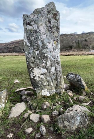 a very tall stone pillar surrounded by smaller stones in the grass. artificial cut marks litter its surface along with moss. behind is a hill and blue sky with clouds