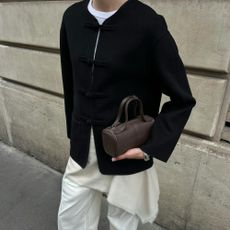 Woman wears black jacket, cream top, white trousers and brown bag 