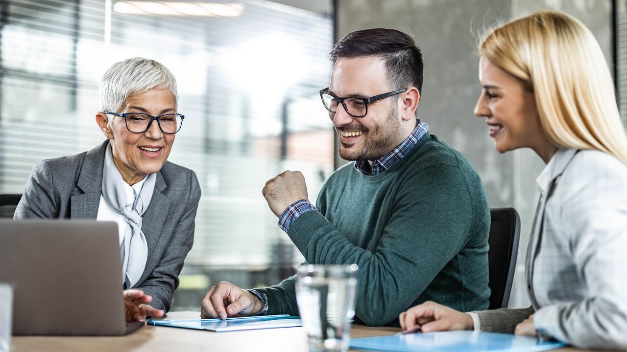 A financial adviser and a couple smile as they look at a laptop together in a conference room.