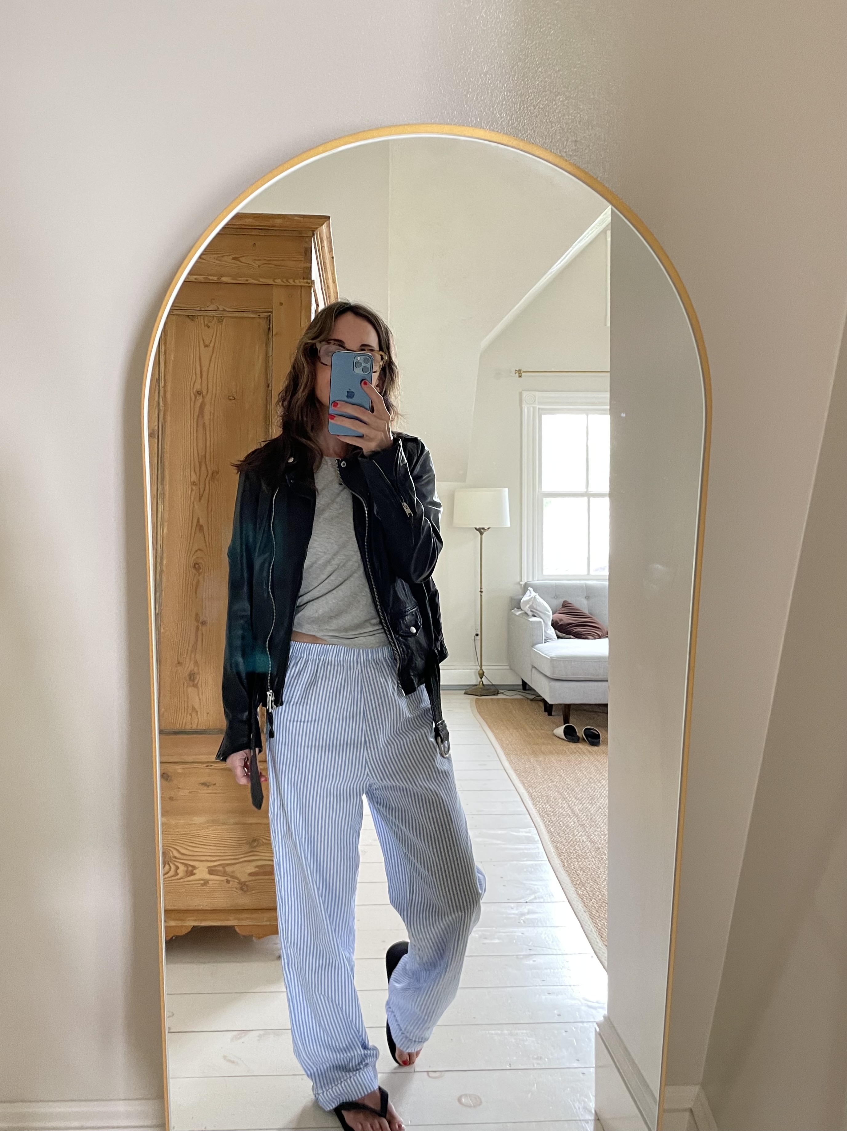 Larissa Mills wearing blue and white striped pajama pants, grey long sleeve shirt, black leather jacket and flip flops in a mirror selfie.