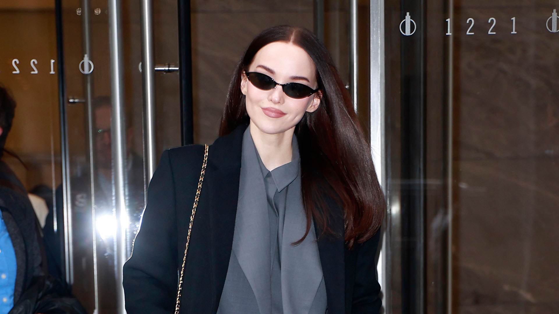 Dove Cameron wearing a black coat, gray top, gray pants, and black heels.