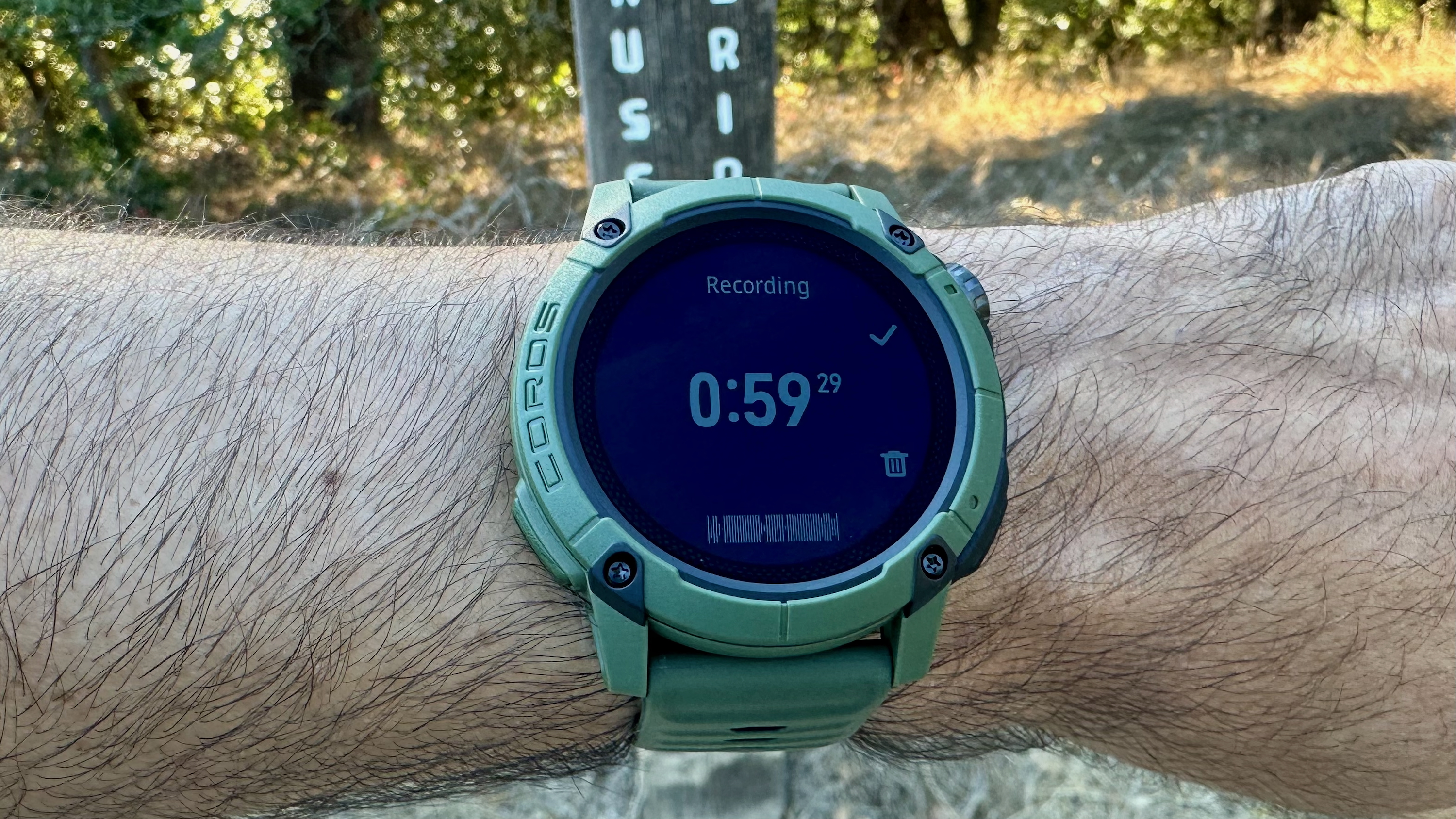 The COROS NOMAD watch showing a "Recording" screen for a voice pin, with a trailhead visible behind it. 