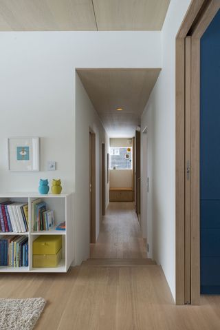 Interior view of the hallway at the Oslo family house featuring white walls, wood flooring, wooden steps, a wood panelled ceiling with spotlights and a window. There is a partial view of a living area