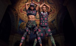 A male and female model standing next to each other with legs apart and arms over their head wearing floral patterned sports clothing.