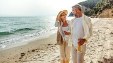 An older couple smile as they walk along the beach with champagne.