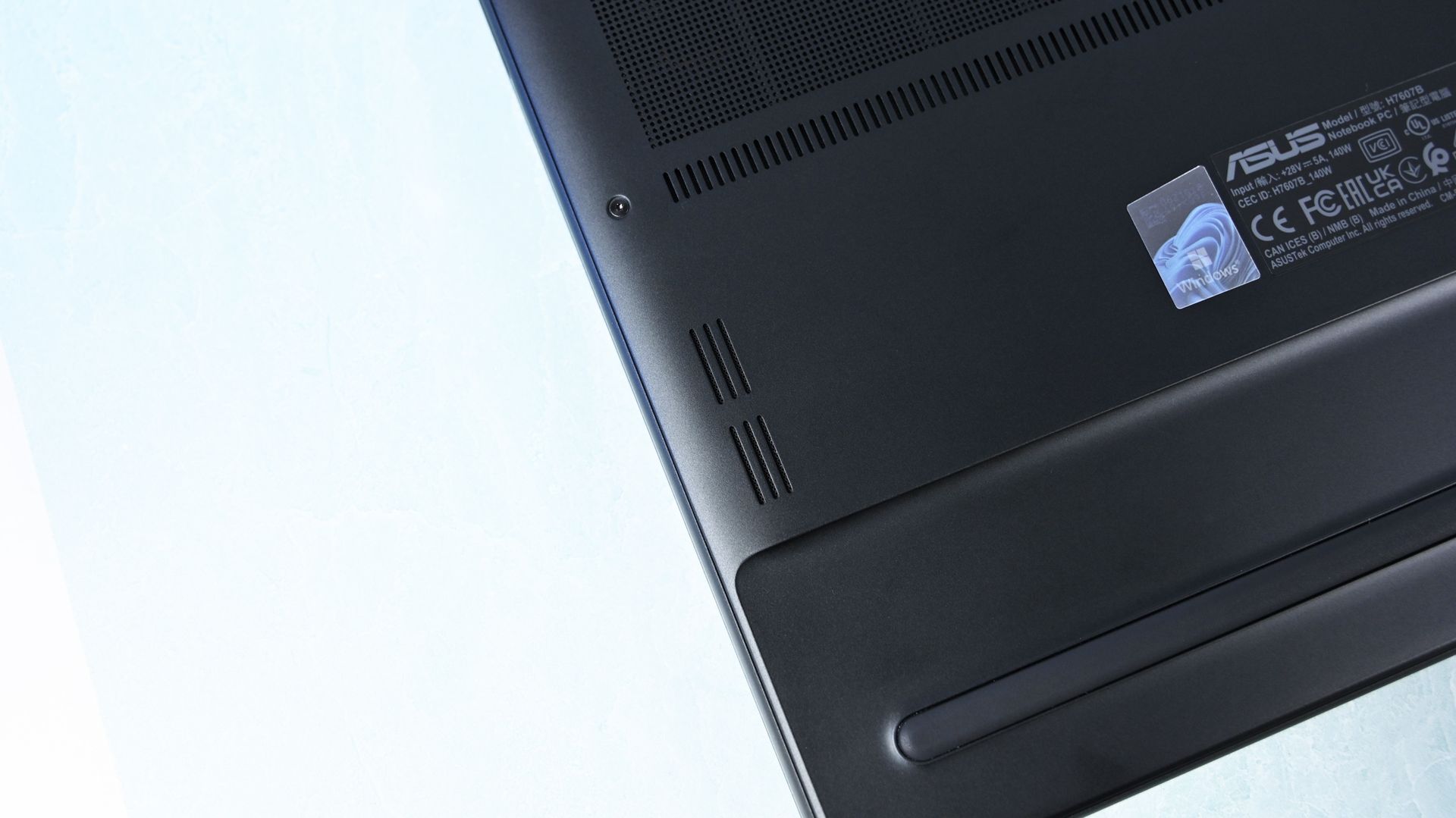 Close-up of the laptop underside corner showing downward-firing speaker cutouts, a rubber foot strip, and product certification labels.