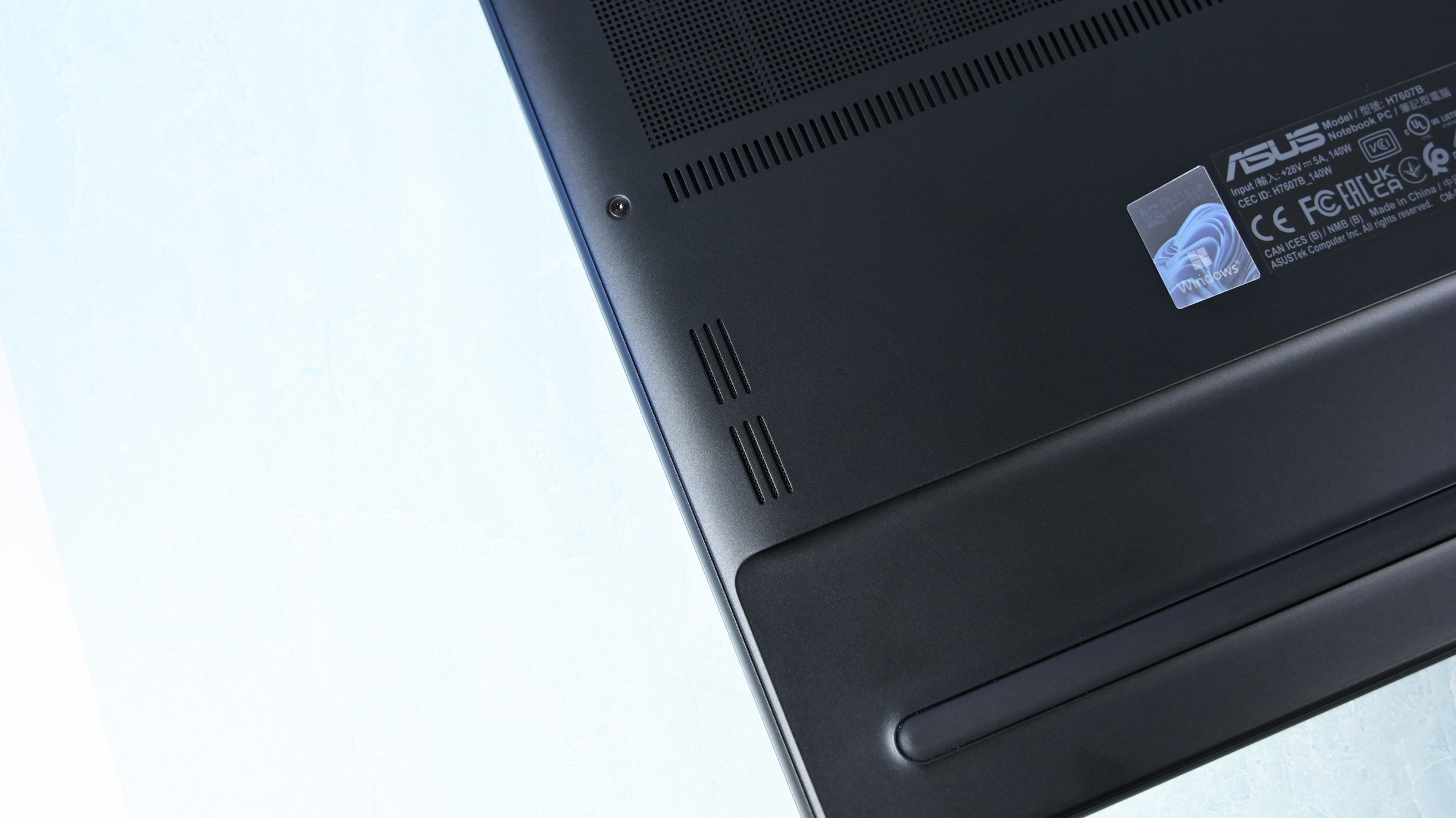 Close-up of the laptop underside corner showing downward-firing speaker cutouts, a rubber foot strip, and product certification labels.