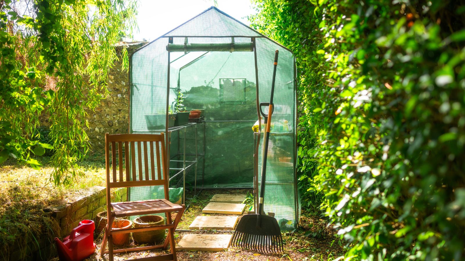 Green mesh greenhouse next to wooden chair and rake