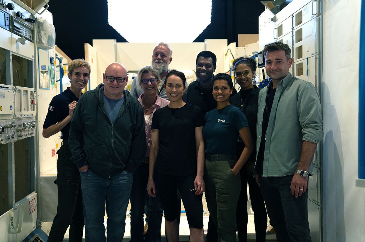 nine smiling people stand in a poorly lit, cluttered hallway