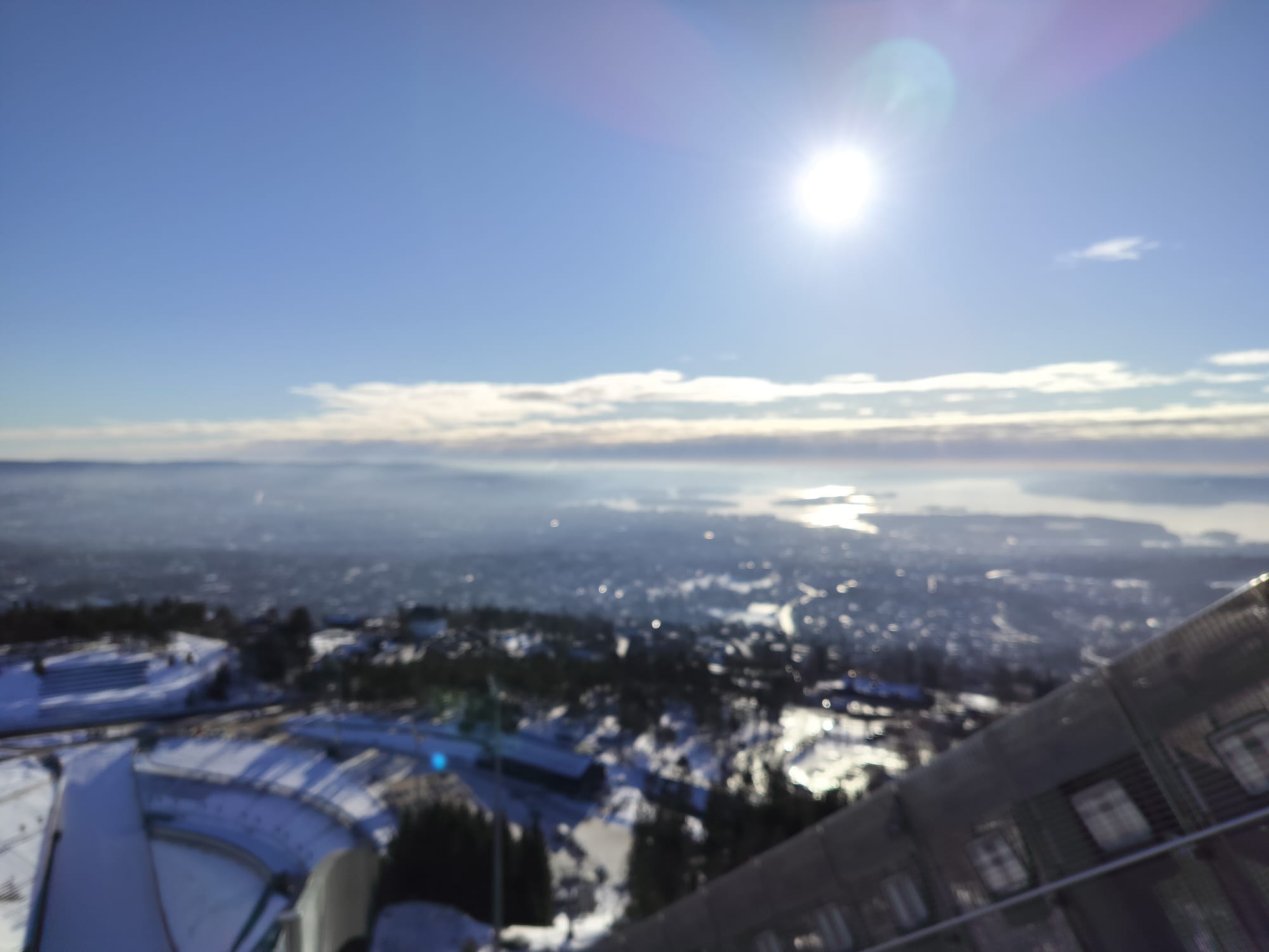 A blurry aerial view from a ski jump tower looks down over a snow-covered city and a hazy lake in the distance.