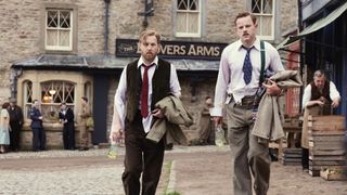 Samuel West in a shirt and waistcoat looking dishevelled and holding a bottle as Siegfried and Callum Woodhouse in a rumpled shirt holding a jacket as Tristan in All Creatures Great and Small.