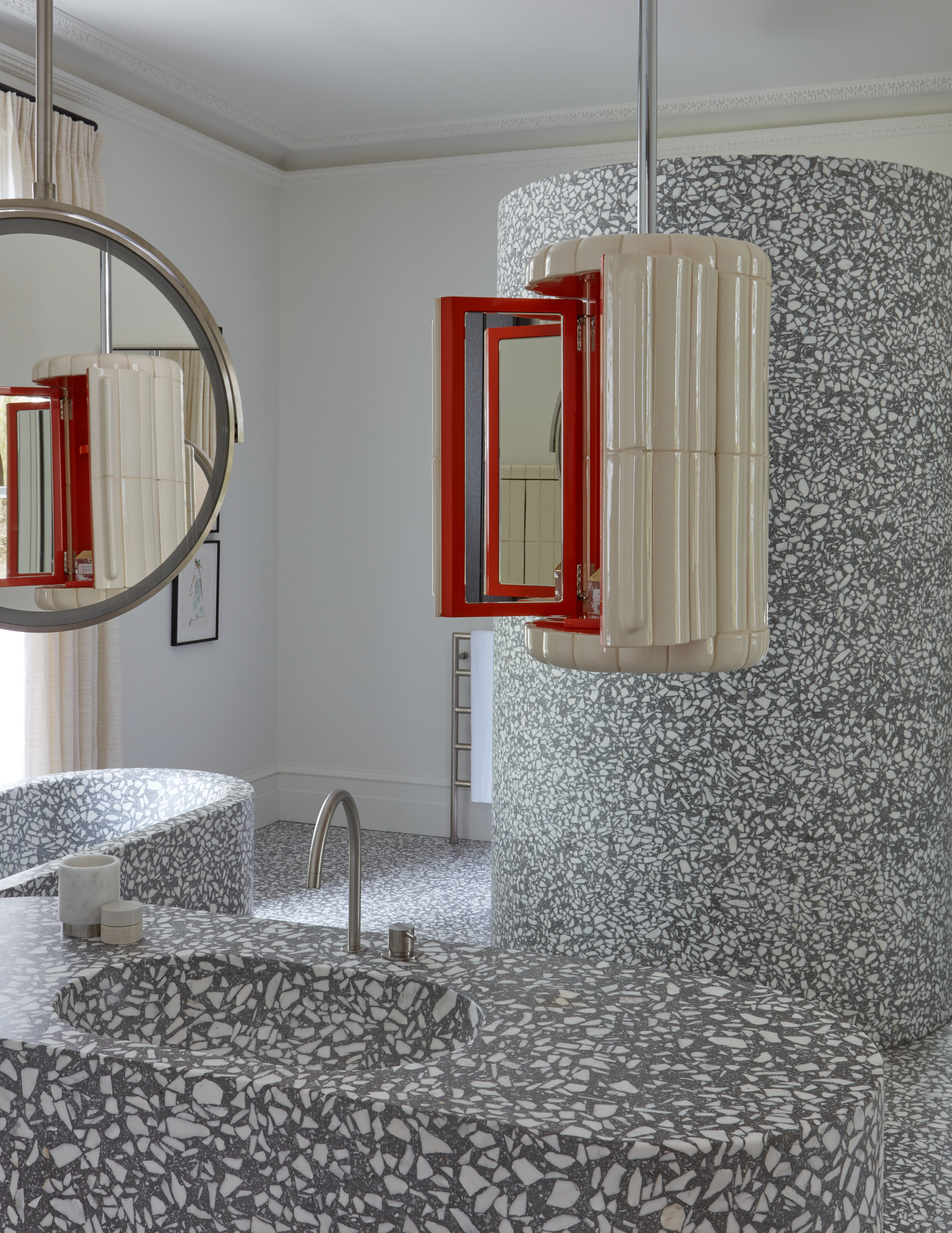Image of a modern bathroom covered in gray and white terrazzo flooring that extends onto the features of the room. Two suspended mirrors are hanging from the ceiling. One is a fluted cream and red material and one is a rounded mirror.