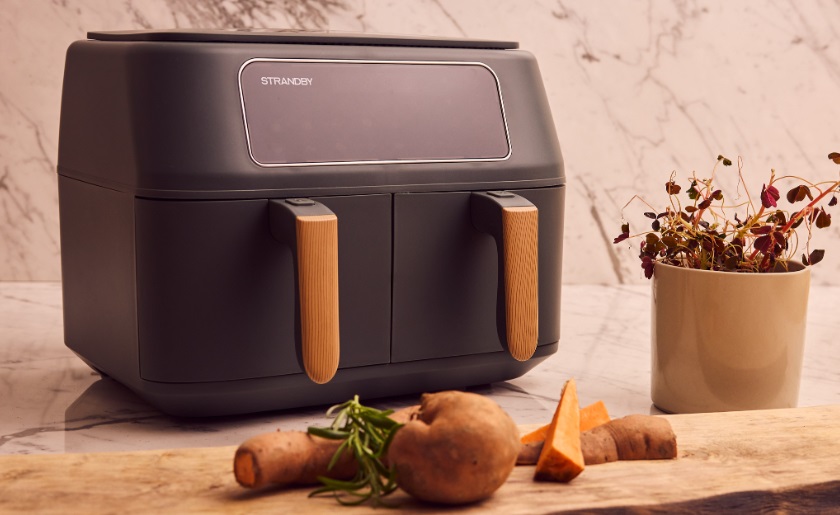 En svart Strandby airfryer står på en köksbänk framför ett par råvaror och en kruka med örter.