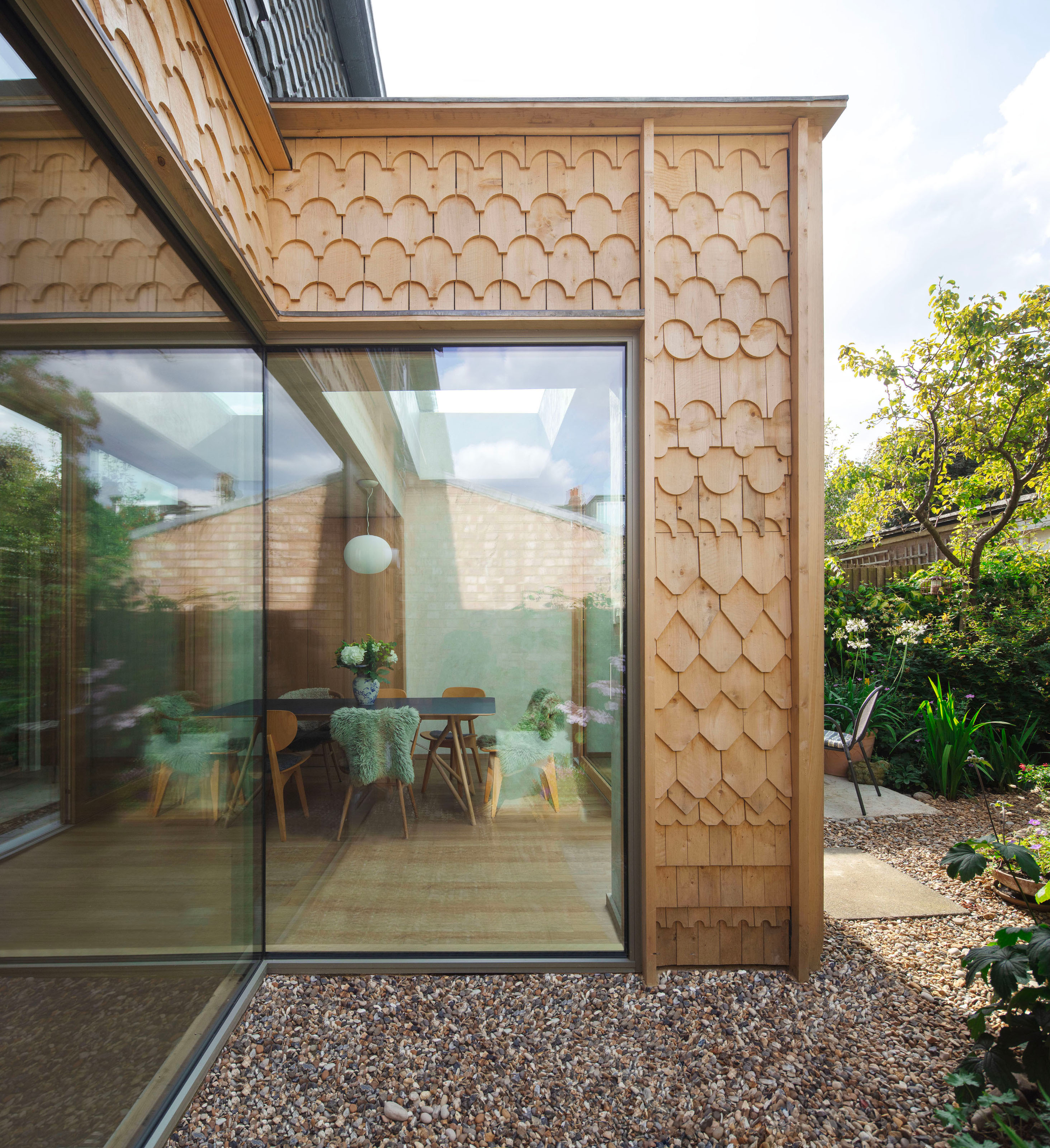 An extension with wood geometric tiling surrounding the full pane windows looking into the kitchen-dining area with gravel surrounding the windows in the outside space