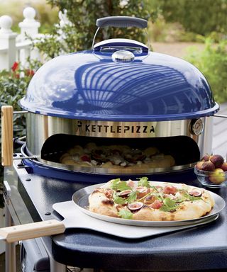 A navy blue kettle pizza deluxe outdoor pizza oven with finished pizza on pizza peel