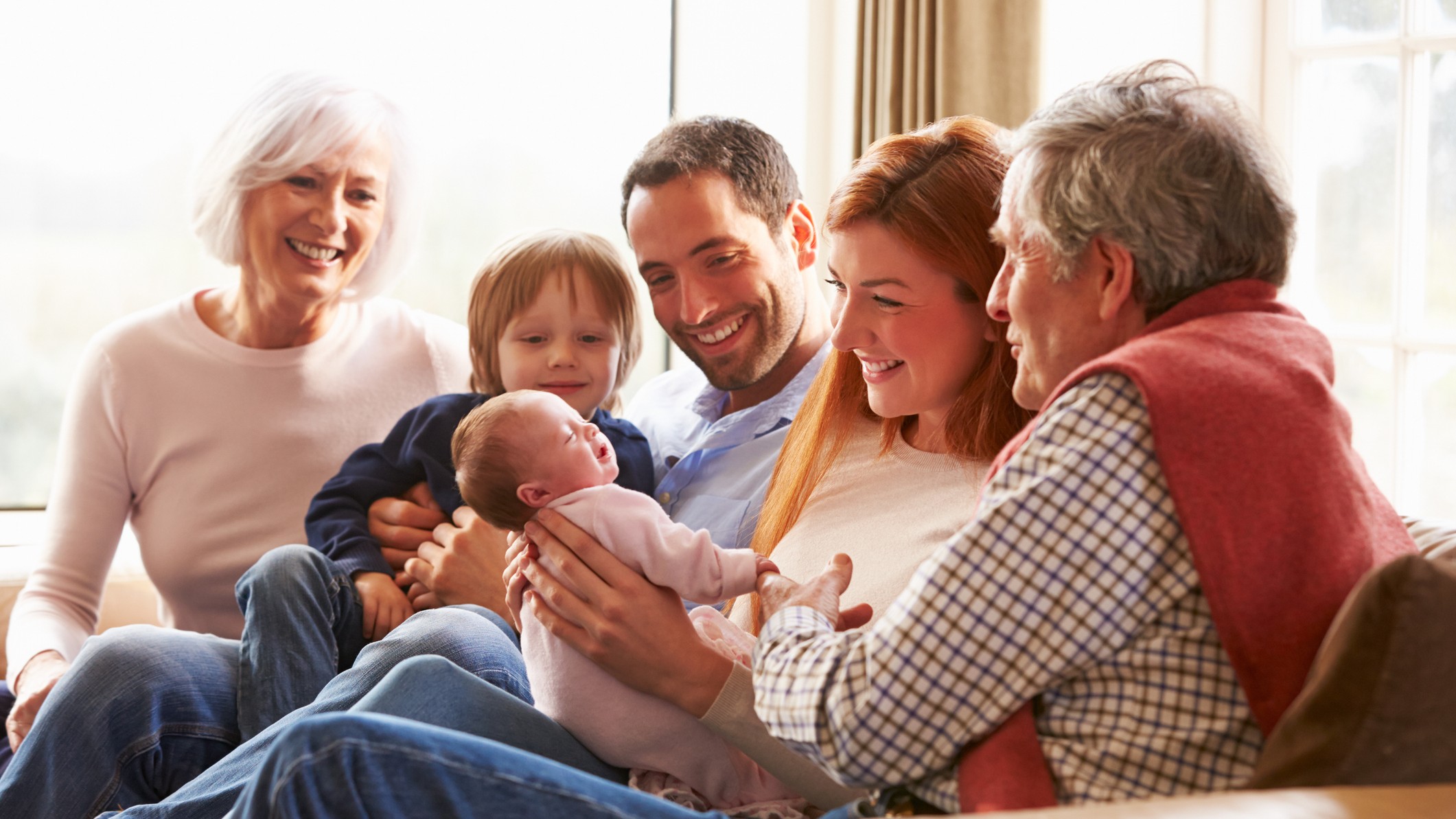 Multi Generation Family Sitting On Sofa With Newborn Baby Smiling.