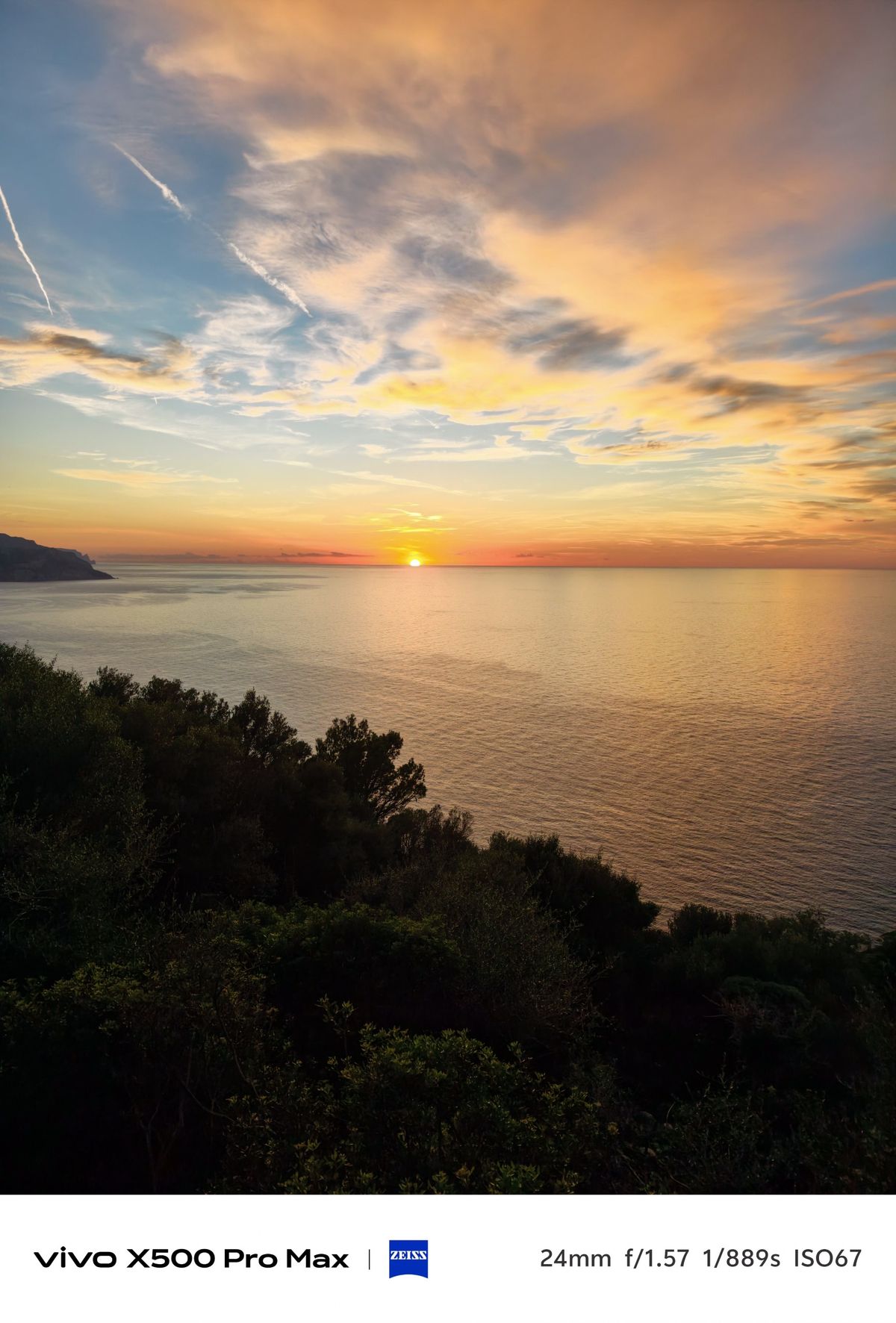 Sunset over a calm sea with orange clouds and dark coastal vegetation in the foreground.