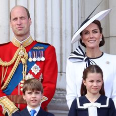 Prince William wears military uniform, Kate Middleton wears a white dress with navy detailing and a matching hat, Prince George and Prince Louis wear matching suits, and Princess Charlotte wears a navy and white nautical-inspired dress