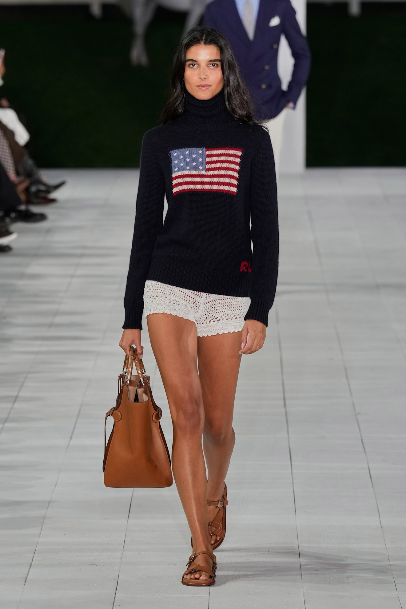 A model at the Ralph Lauren show during S/S 25 fashion week wearing a loose-knit cotton sweater with an American flag on it with shorts and sandals.