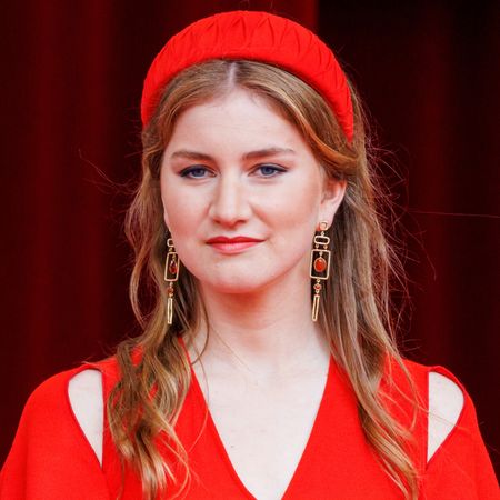 Princess Elisabeth of Belgium wearing a red cold shoulder dress, dangling red and gold earrings and a puffy red headband smiling at the camera