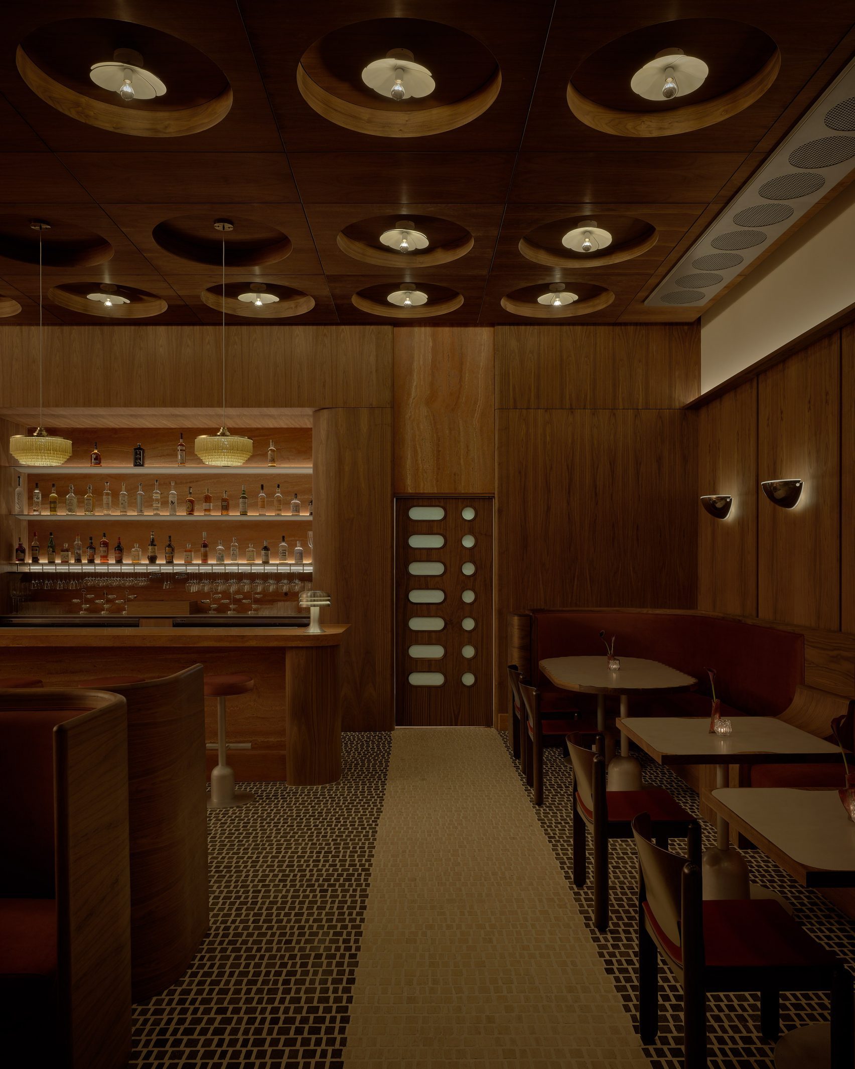A dimly lit living room with tiled checkered floors in cream and brown, pale brown and terracotta velvet furnishings, pendant chandeliers in pale gold, and spherical ceiling lights and wall sconces.