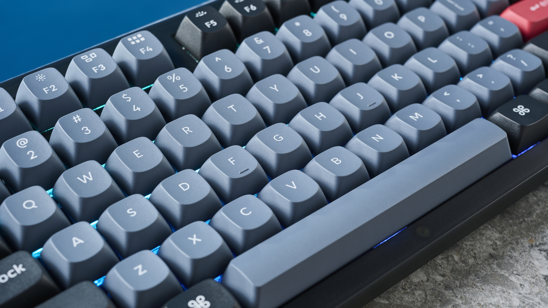 A photograph of the Keychron V6 in black, with gray , black and orange keycaps. The keyboard is positioned on a stone slate, with a blue wall in the background.