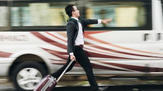 A man wearing a suit runs after a bus with a suitcase.