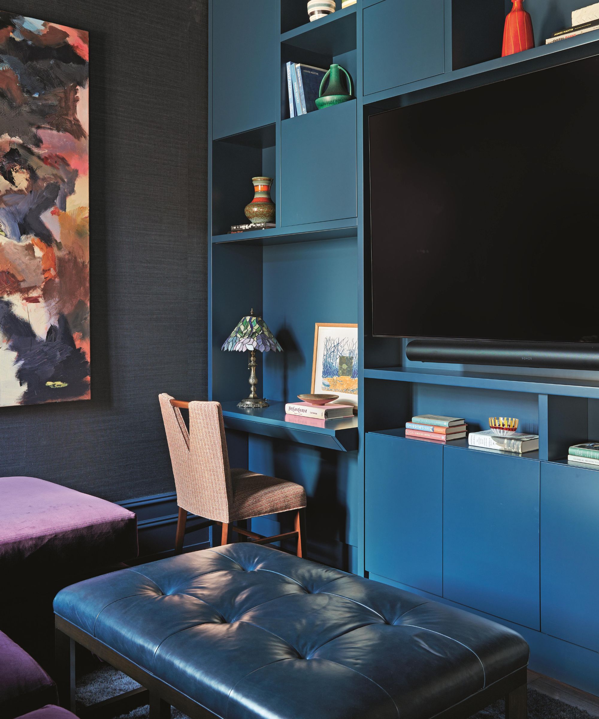a cobalt blue TV and media room in a new york apartment with a built in bookcase and storage system around a large TV with a small desk and large artwork
