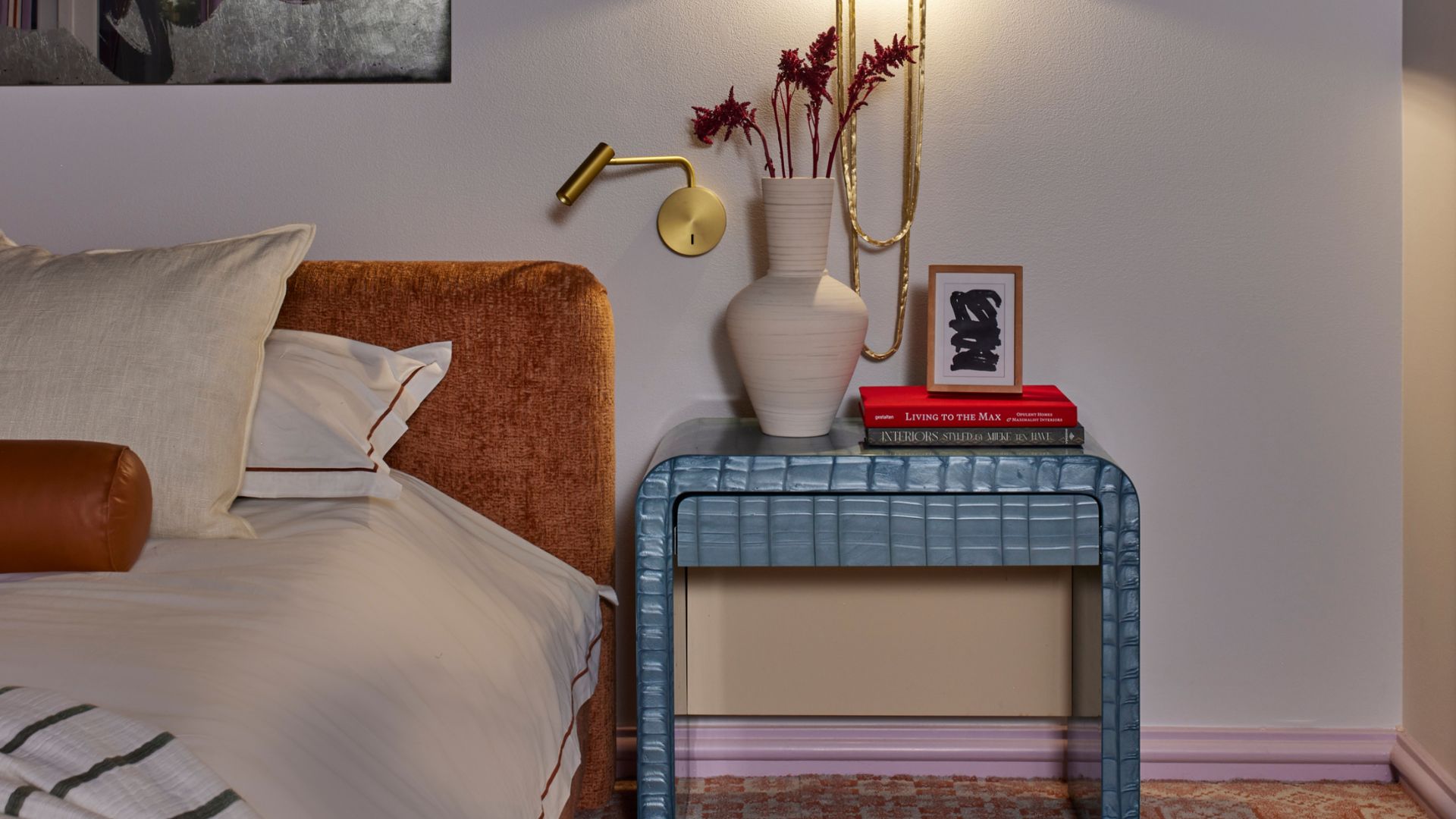 A white bedroom with a blue tiled nightstand with a white vase, a stack of books, and a lamp sconce above