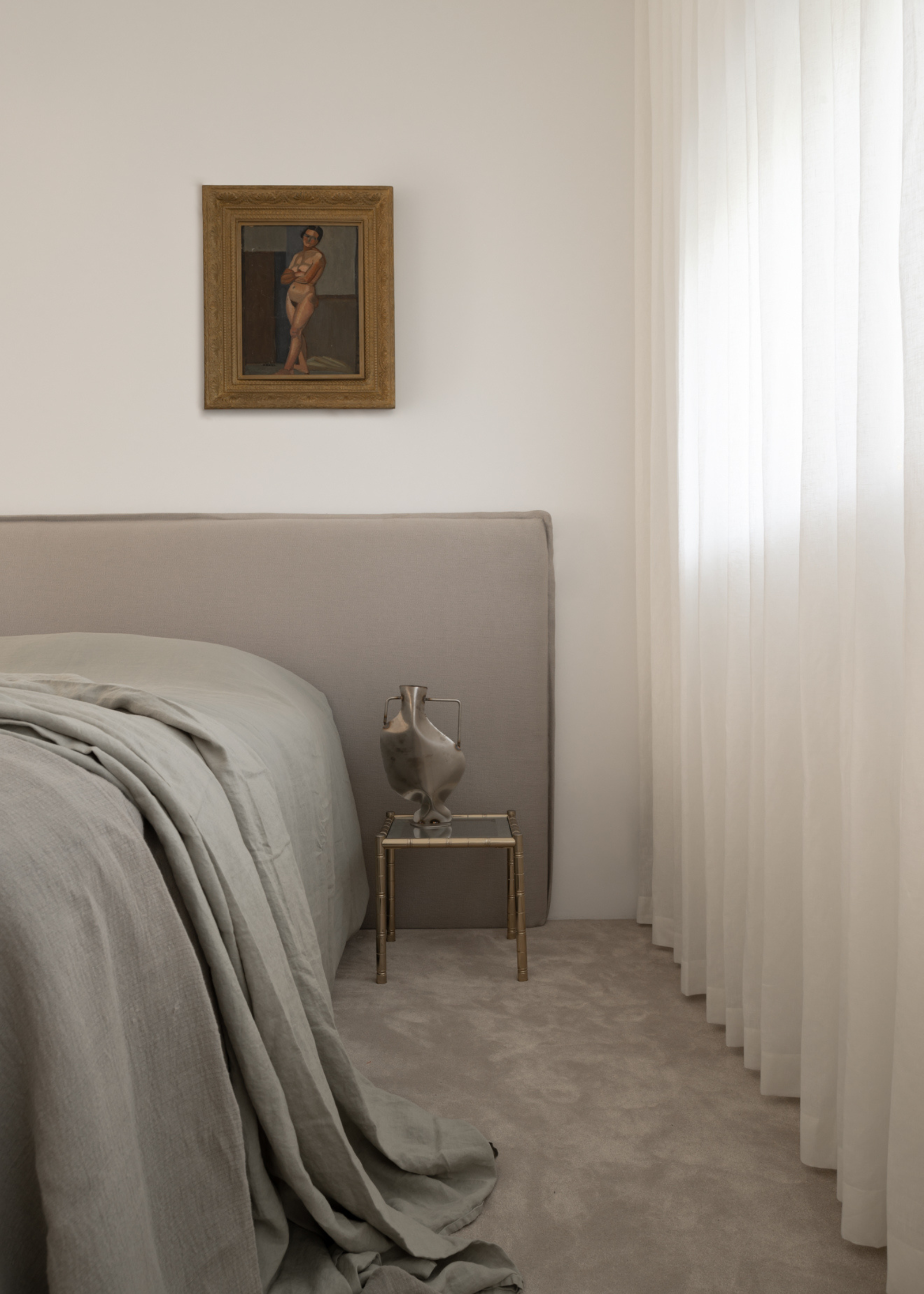 A white bedroom with sheer curtains, a glass side table with a silver vase, a framed painting above the bed and gray sheets