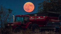 An orange full moon is photographed above a red tractor in a field at night.