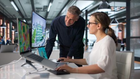 BIM Manager And Team Review 3D City Model Using Laptop Software. An older business owner looks at a monitor with a younger worker in a modern office.